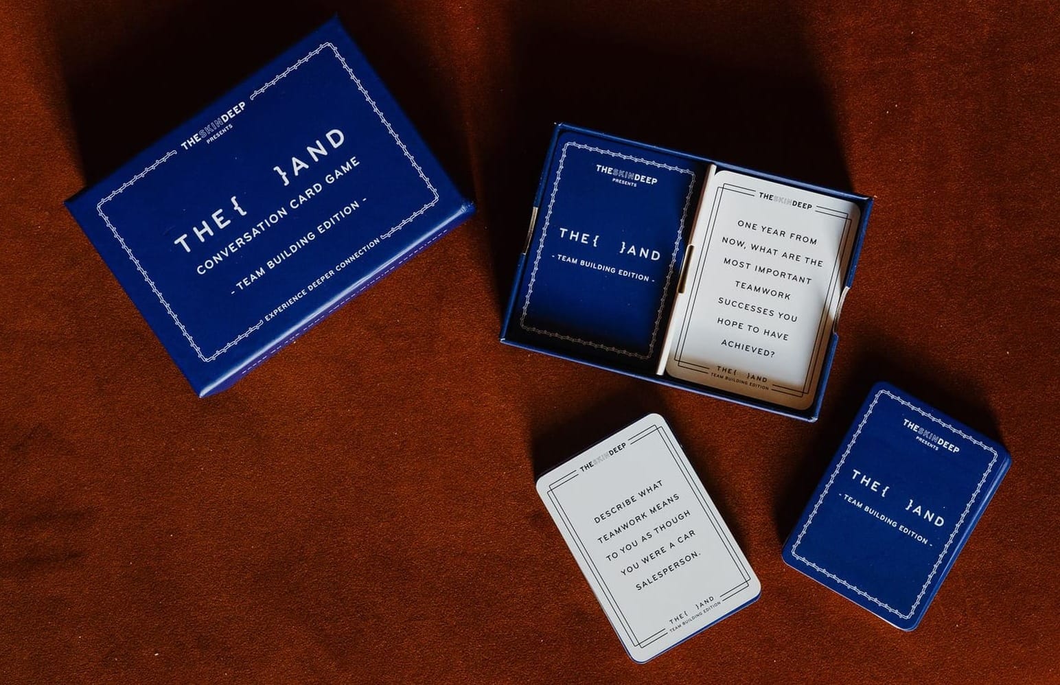 {THE AND} Team Building Edition box and cards laid out on a rust-colored surface, with two question cards face up.