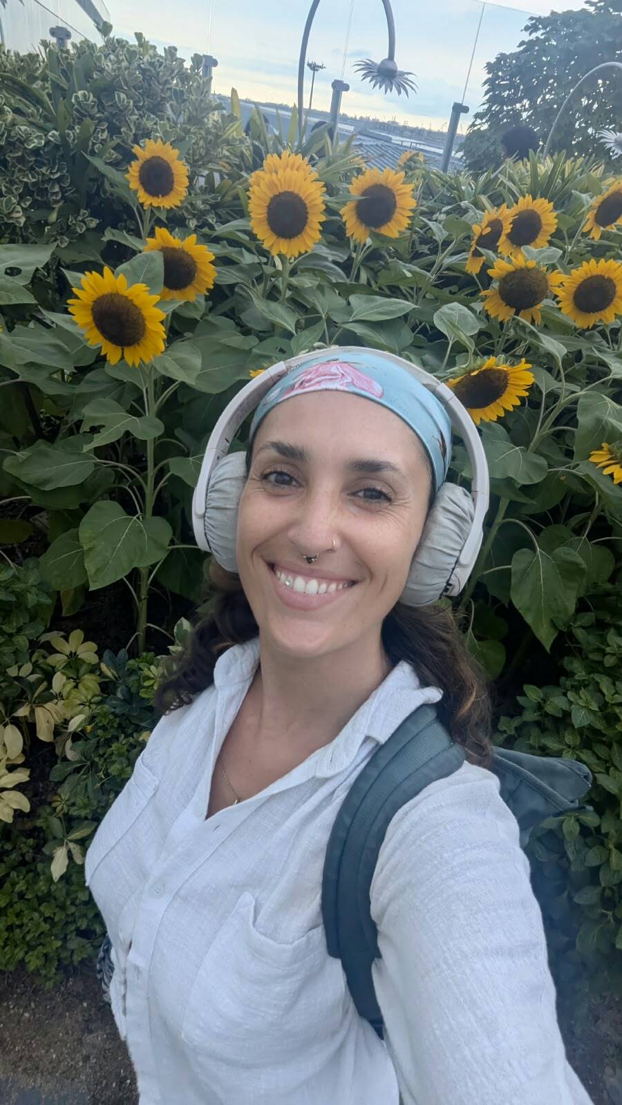 Gianna Shamone smiling in a selfie, headphones around her neck, standing in front of a bed of sunflowers.