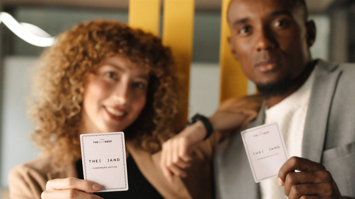 Two coworkers holding up  {THE AND} Coworkers Edition cards and looking at the camera