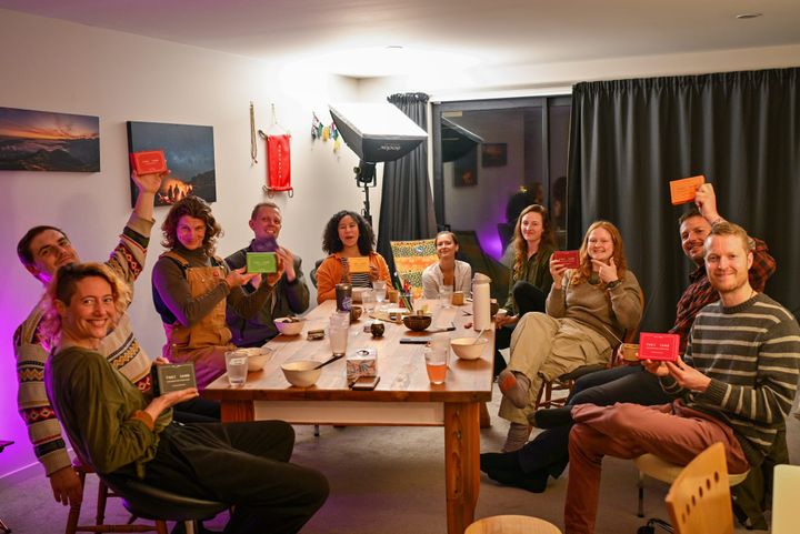 A group of ten gathered around a table in Ōtautahi Christchurch, several holding up colorful {THE AND} card decks, smiling for the camera.