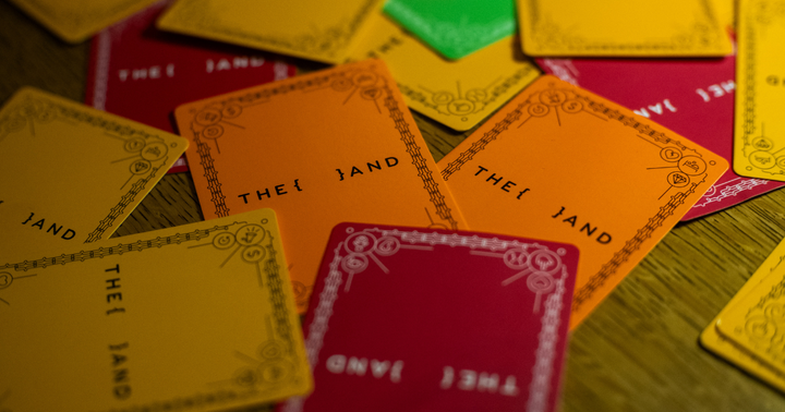 Close-up of scattered {THE AND} conversation cards in yellow, orange, red, and green, spread face-down across a wooden table.