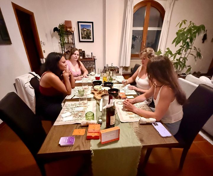 Four women gathered around a dining table at Missy's home in Sacile, Italy, playing {THE AND} card game together over wine and food.