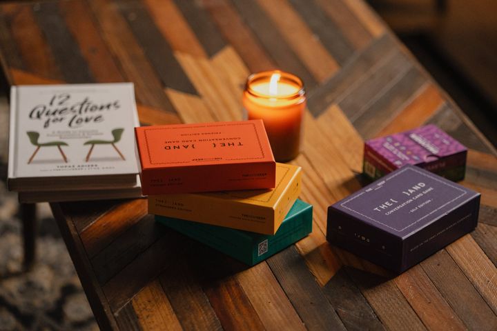 A candlelit display of {THE AND} card game boxes in multiple editions — Friends, Strangers, Self, Family and Kidulting — stacked on a wooden table beside the book "12 Questions for Love."