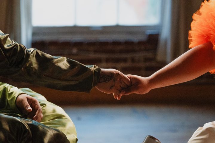Close-up of two people's hands clasped mid-handshake, lit by warm window light. One person wears an olive satin jacket with a visible hand tattoo; the other wears an orange tulle sleeve.