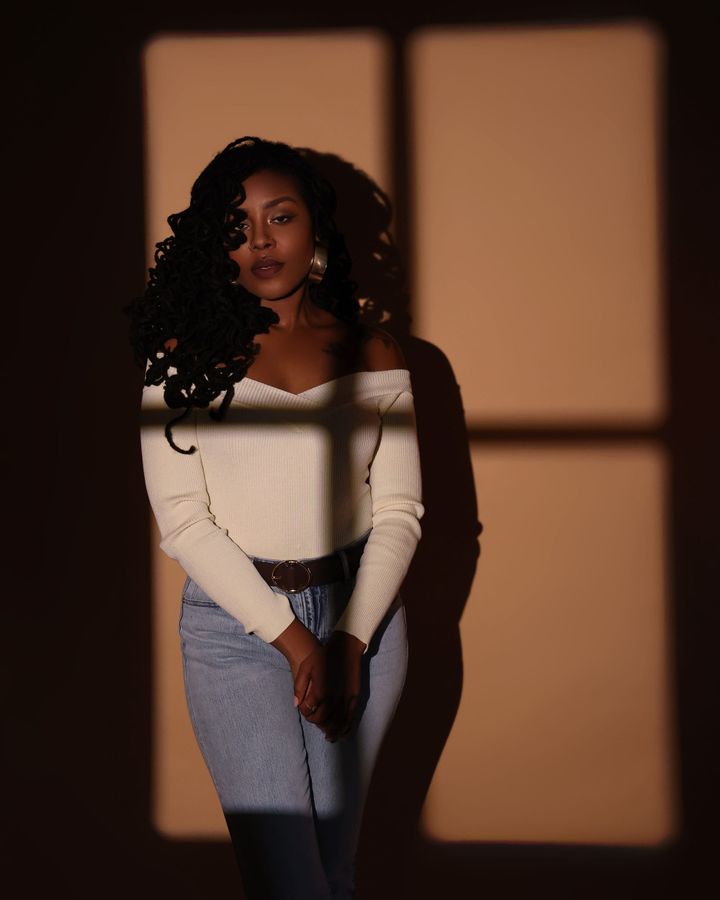 DeJa Jovon Survillion standing against a wall with window-shaped light, in a cream off-the-shoulder top and jeans.