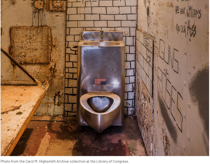 a shiny steal prison toilet in just about the most disgusting dirty peeling paint rusty prison cell you can imagine. The toilet has an upside-down American flag sticker on it