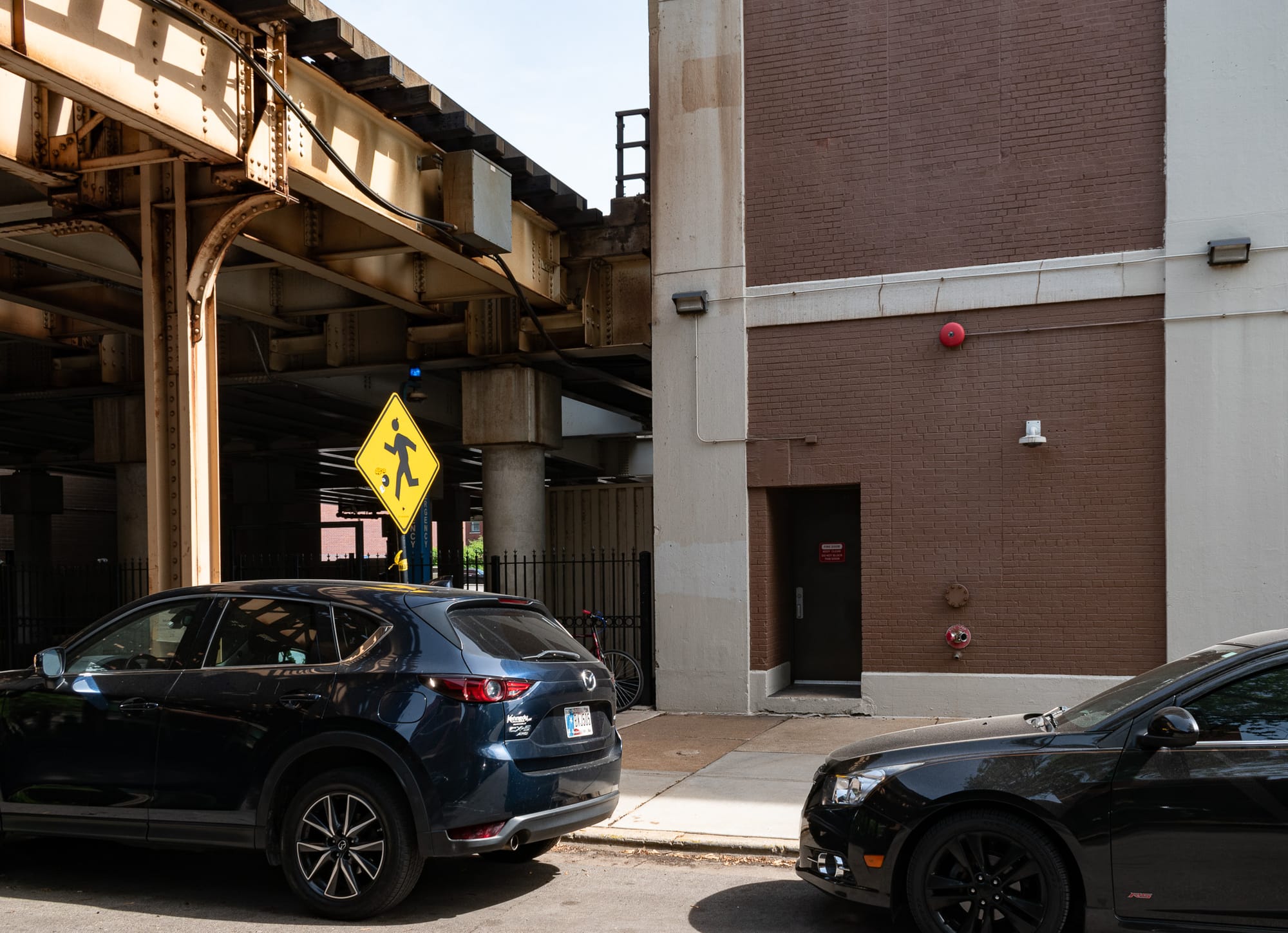 Steel structure of the L on the left, a multi-story masonry building in the center and right. Yellow children at play sign. Two cars. 