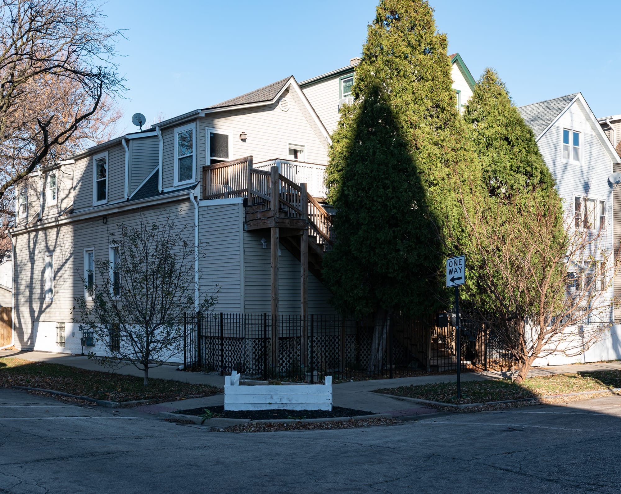 Streetcorner with a beige house on it with big wooden stairs, a white planter box on the corner, a one-way sign, and a very large evergreen tree.