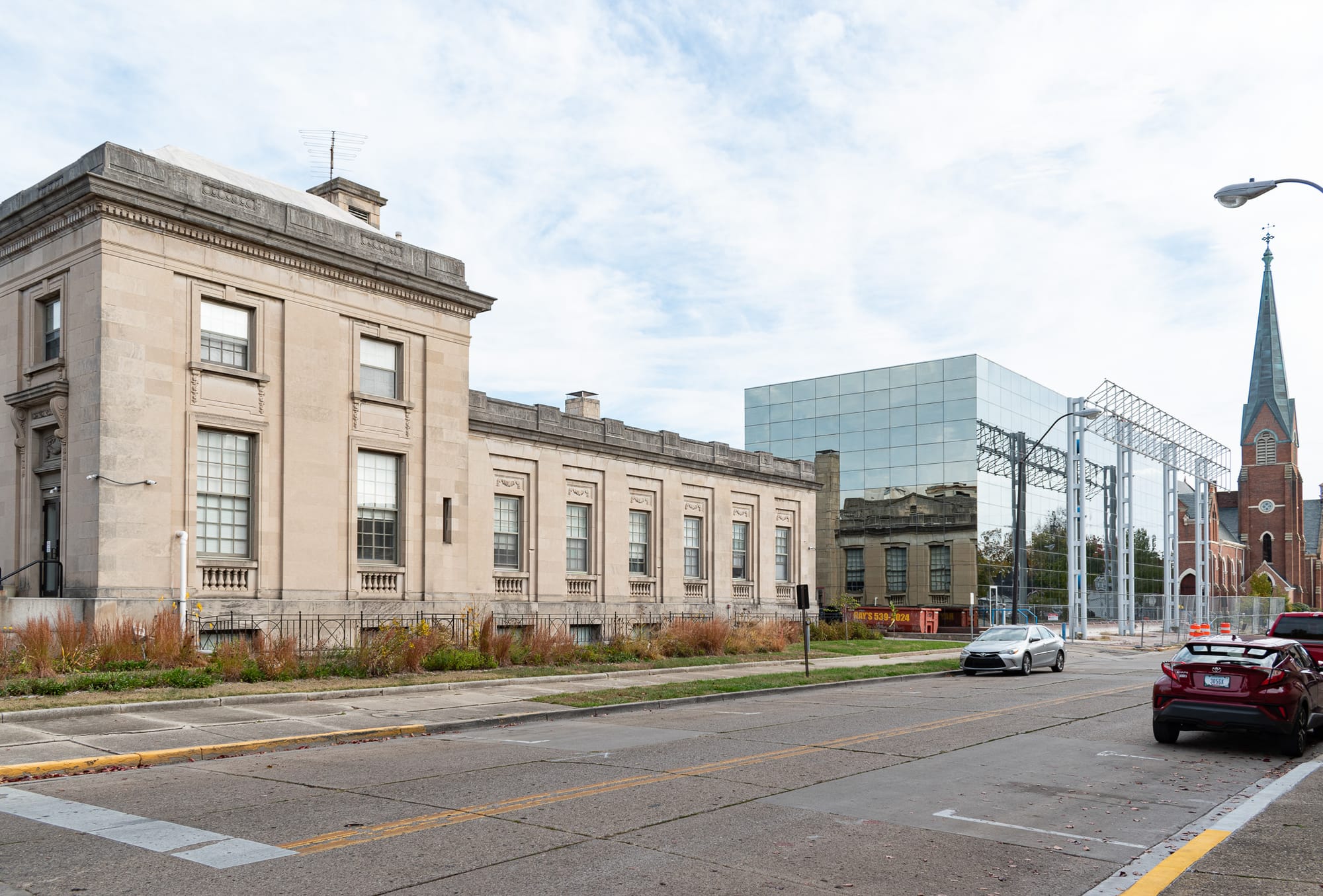 A two story stone former post office building on an asphalt city street with a mirrored glass rectangle next door, you can see the post office reflection in the glass, then a church steeple further down the street.