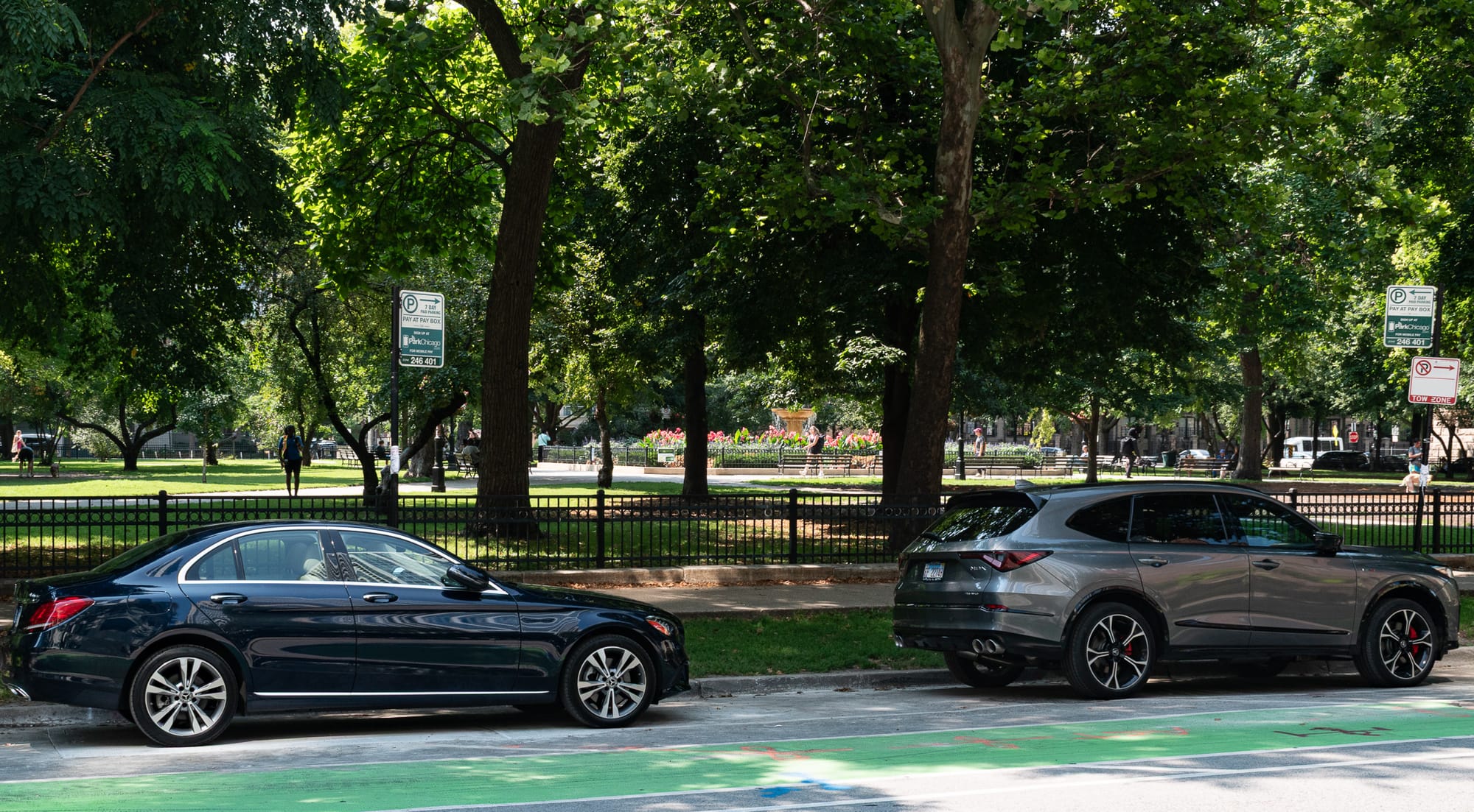 A Chicago street with a green painted bike lane, two parked cars, trees, and the fountain of Washington Square Park in the distance.