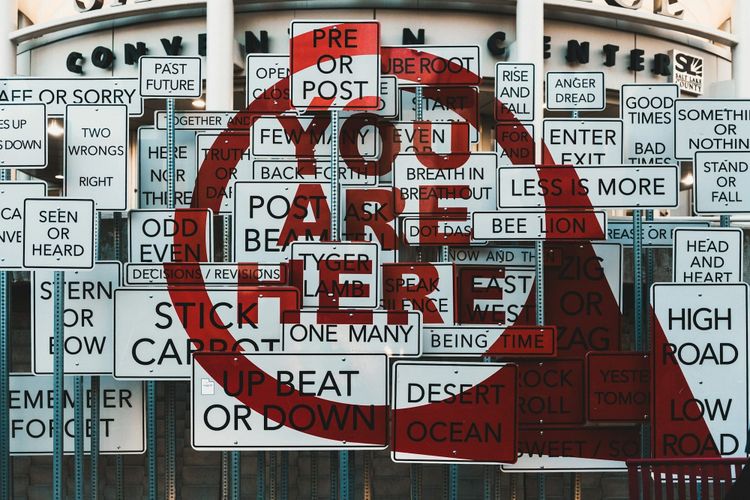 Art installation of contradictory road signs, from Past or Future to Odd or Even, overlaid with a red YOU ARE HERE, echoing catalogue descriptions that all sound authoritative.