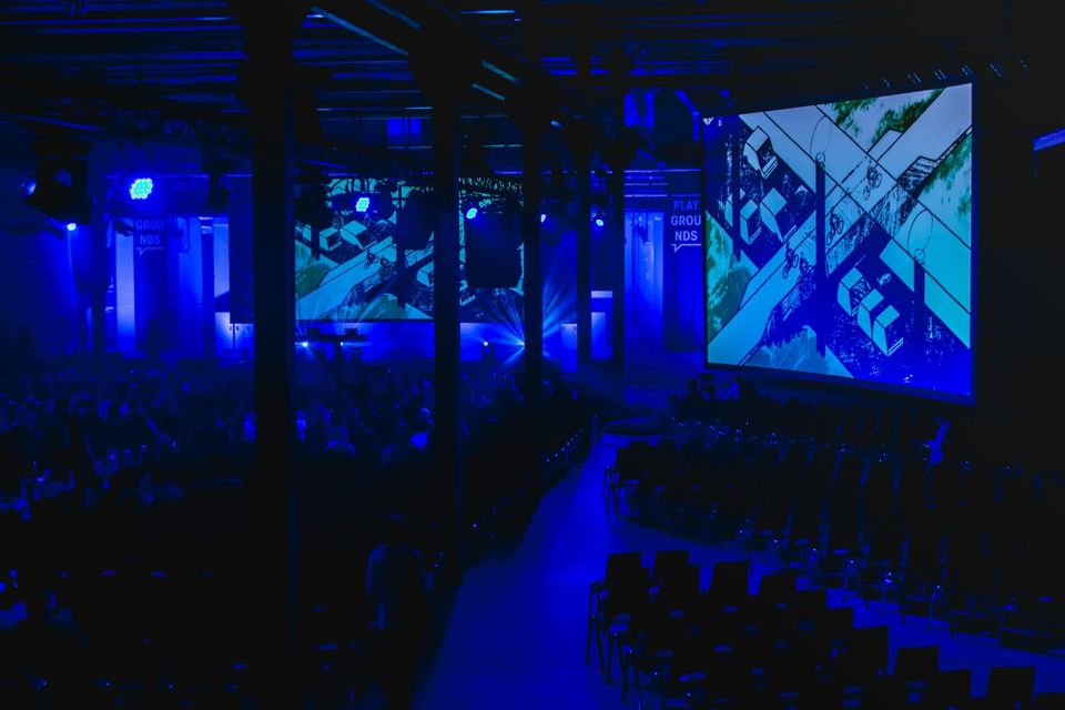 A wide, high-angle view of a large tech conference venue in deep blue tones with a massive screen displaying technical architectural diagrams, representing high-end professional leadership.