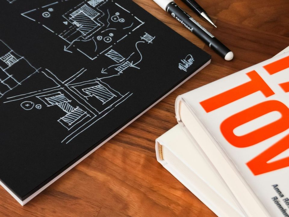 Professional wooden desk with books, architectural sketches on a black notebook, and drafting pens. Represents strategic technical leadership.