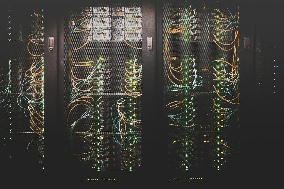 Dark data centre cable network photographed through server rack mesh doors, representing the dense interconnections an AI agent can traverse without a deliberately designed access boundary.