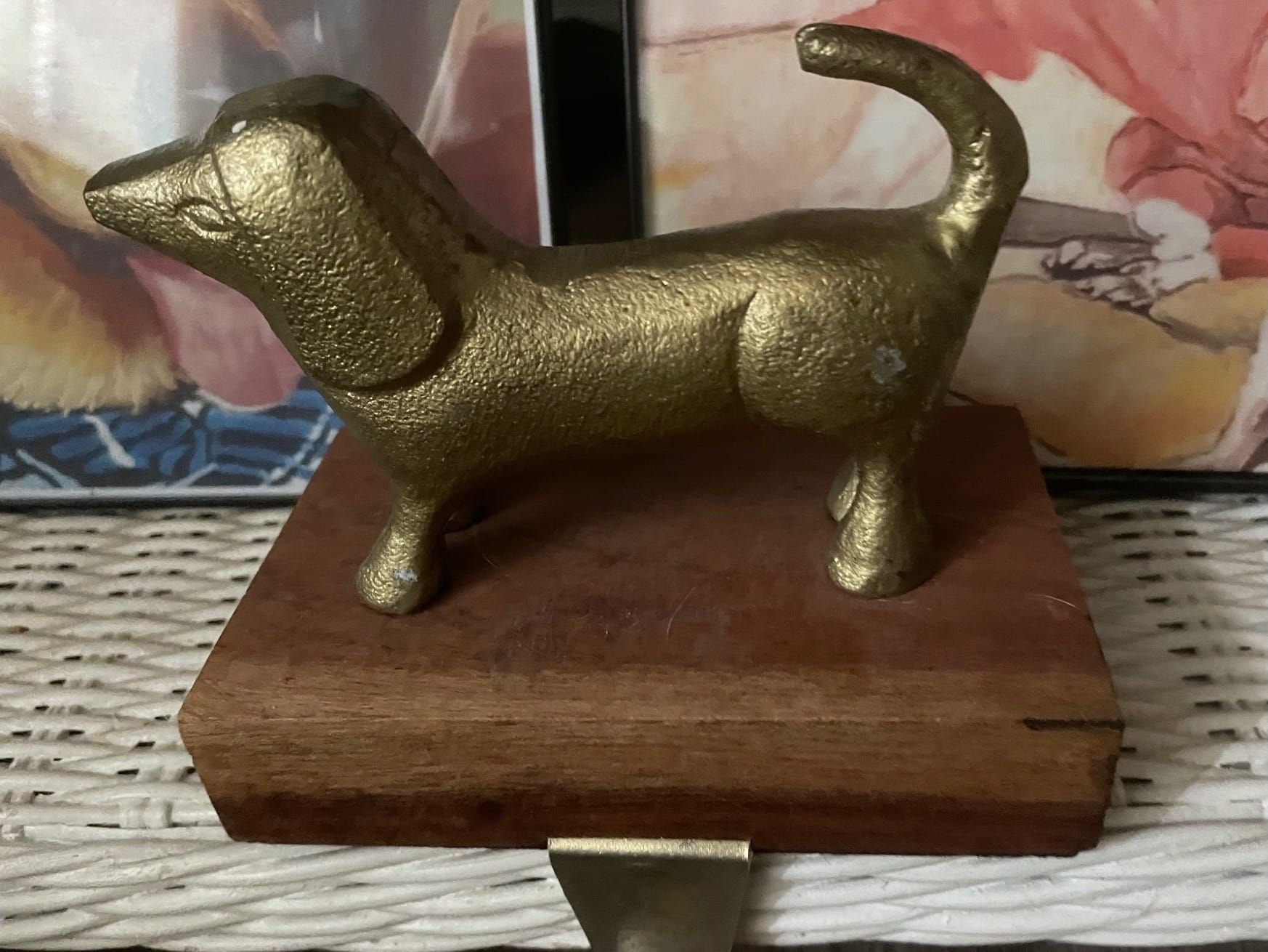 Photo of a golden figurine of a dog mounted on a wooden plaque on a white wicker shelf with framed pictures behind it.