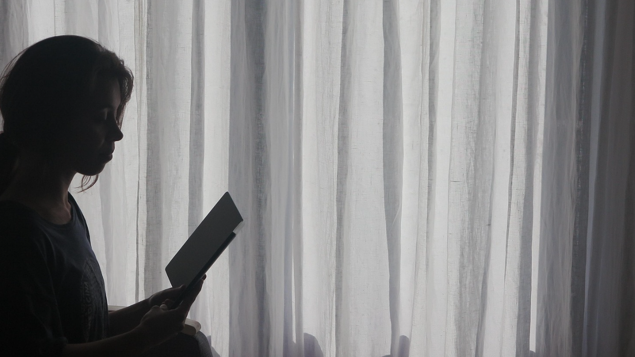 Silhouette of young woman reading her phone in the dark while sitting up in bed, with light streaming in through sheer curtains over the window