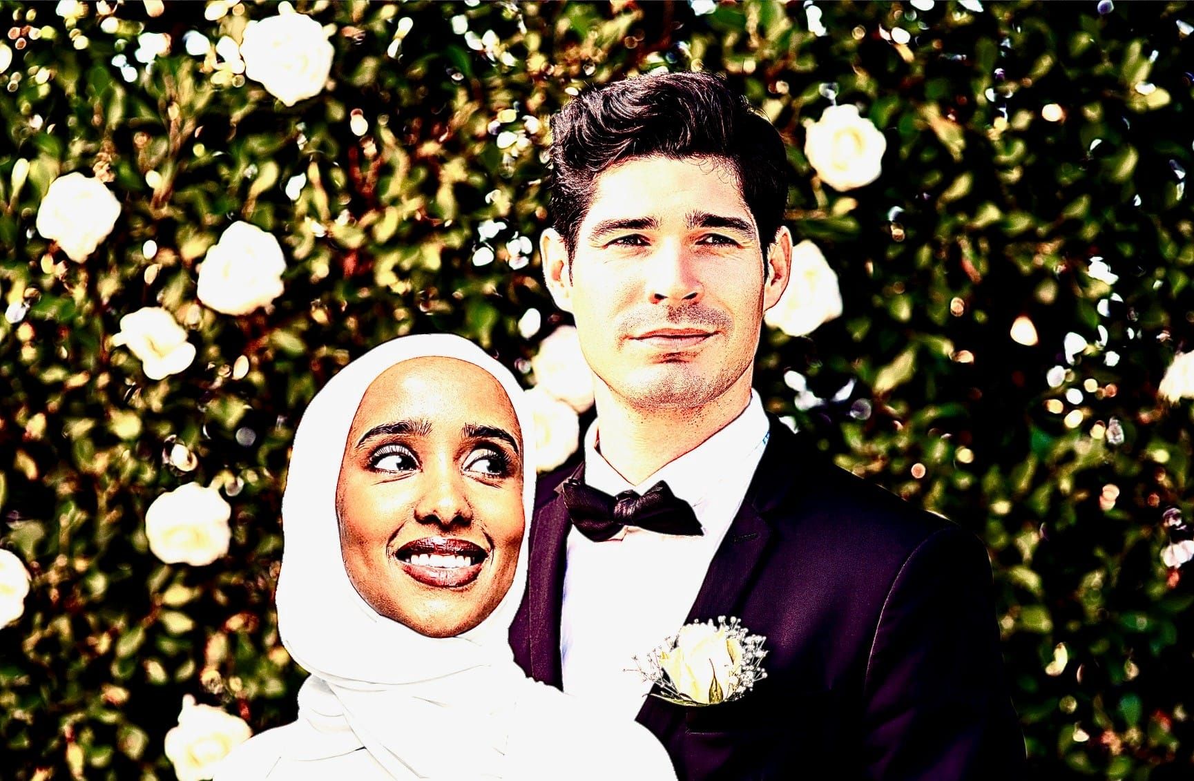 Hispanic man in tuxedo and smiling woman of color in white hijab and gown in front of a rosebush