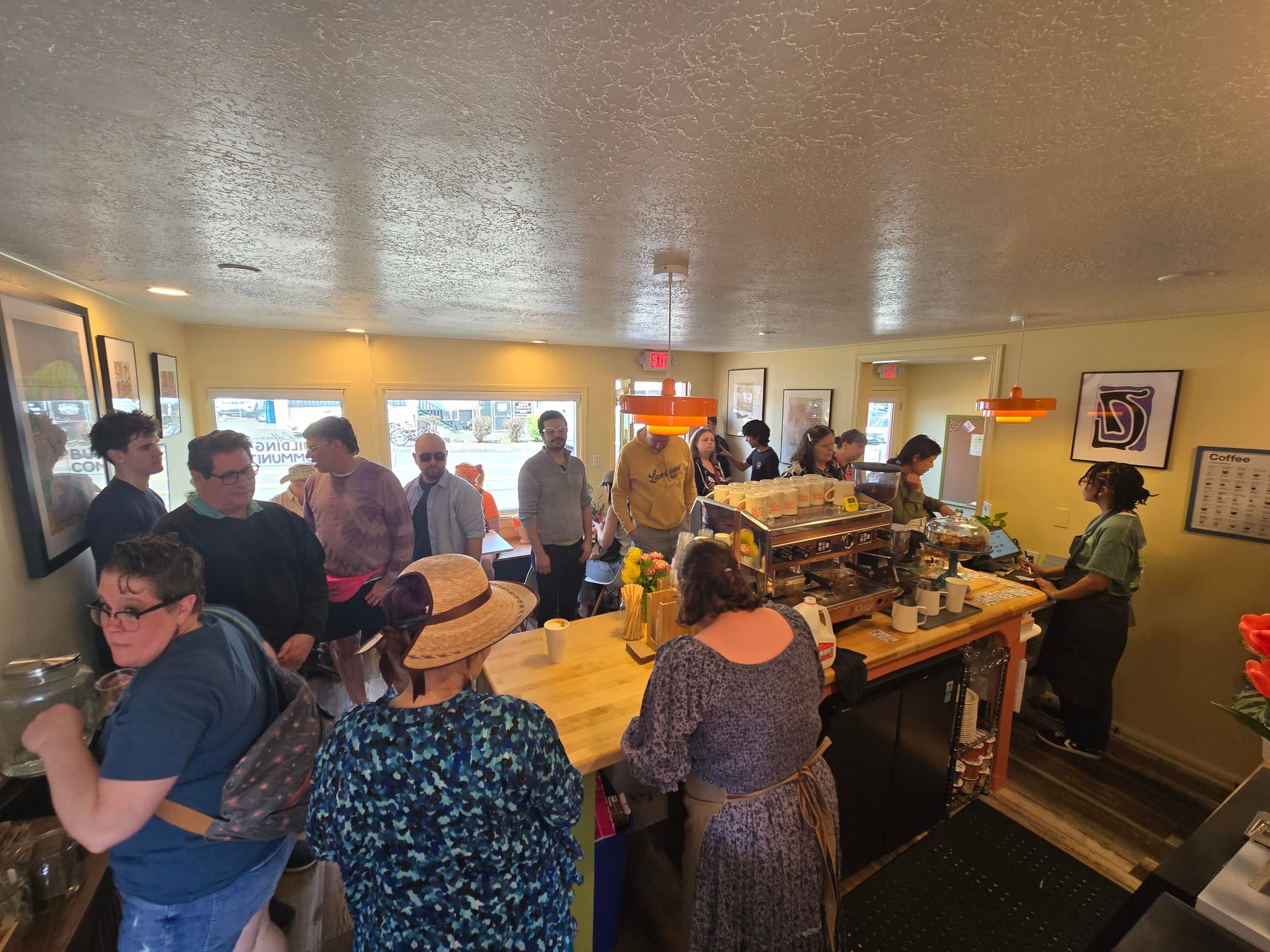 Photo taken from behind the coffee counter, showing over a dozen people standing around waiting to order or receive their coffee drink.