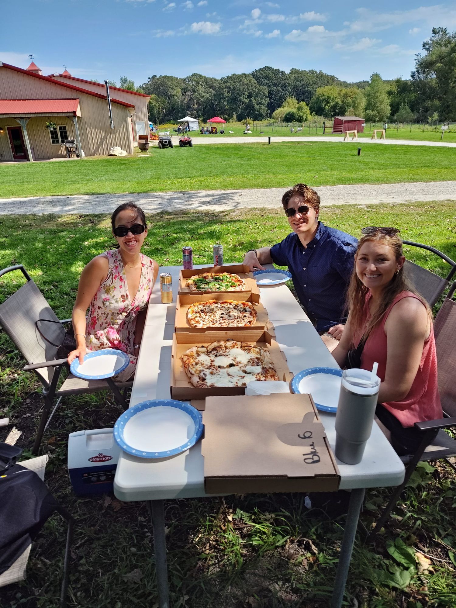 two women and one man smiling and sitting at a table with three pizzas at a farm