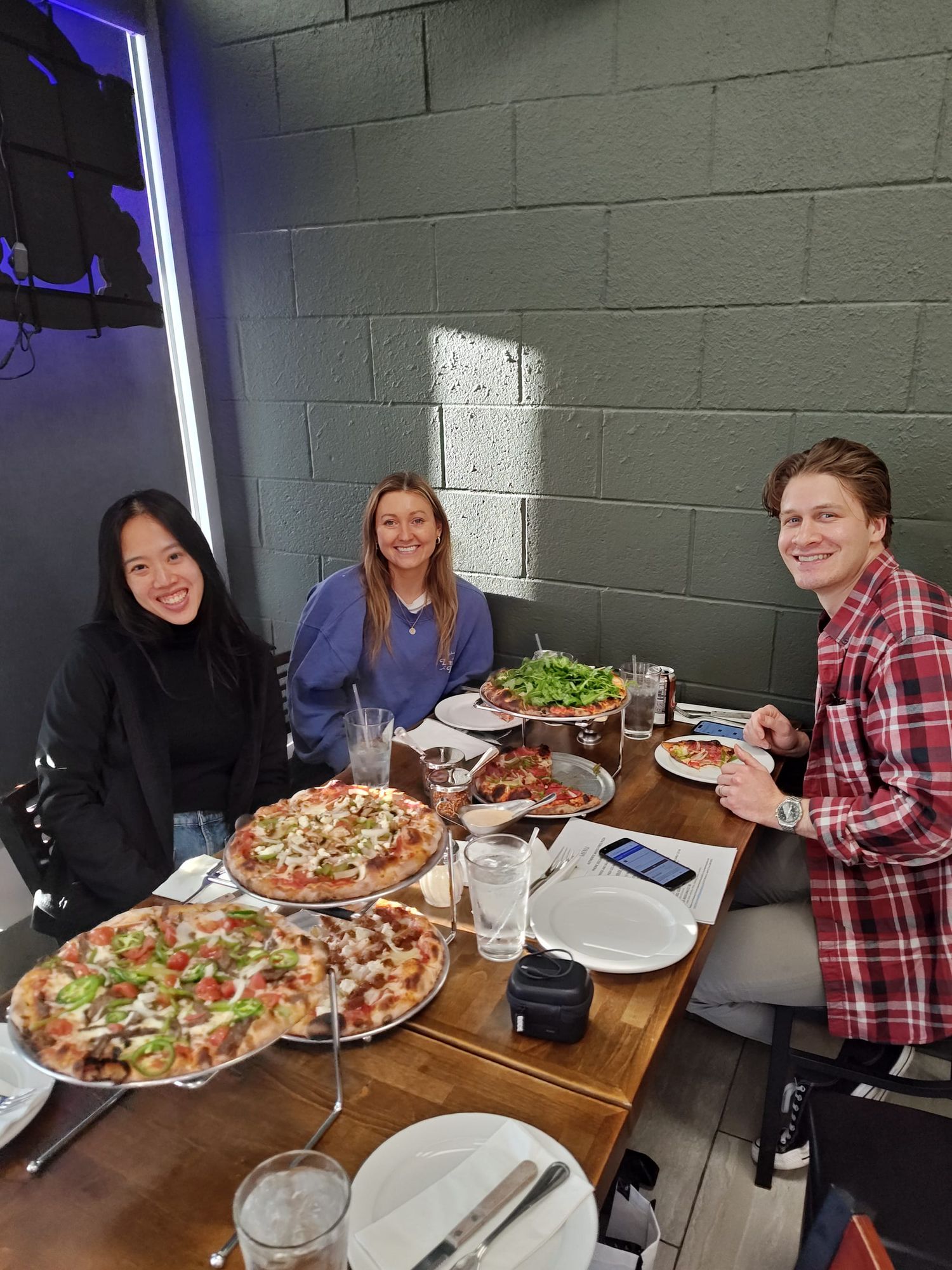 three people smiling and sitting at a table with five pizzas on the table