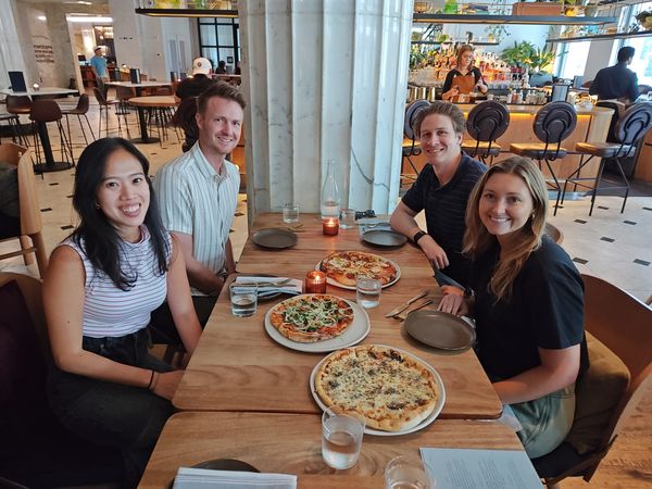 four people smiling and sitting at a table with pizzas on the table