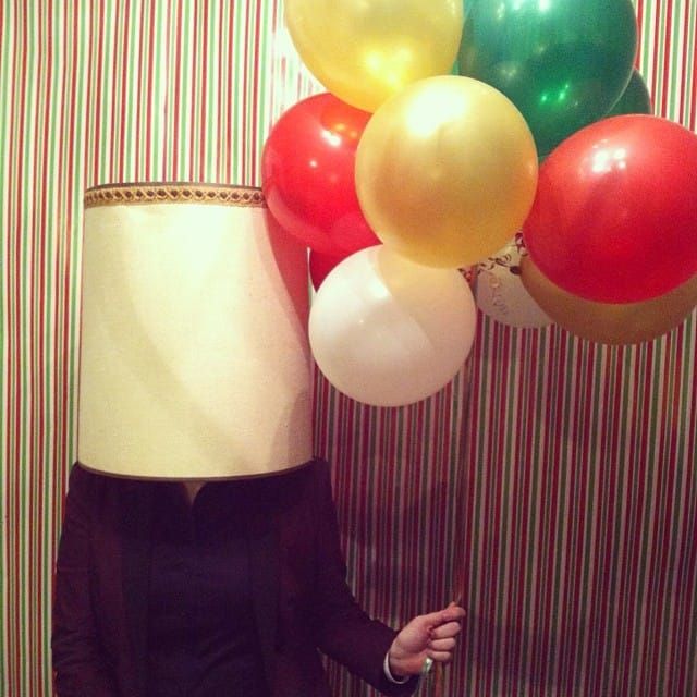 A person with a lampshade completely obscuring their features holding a bunch of balloons in front of a striped background