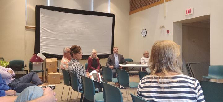 A meeting in the meeting room. Three people visible at the table, and six people seated in the crowd.