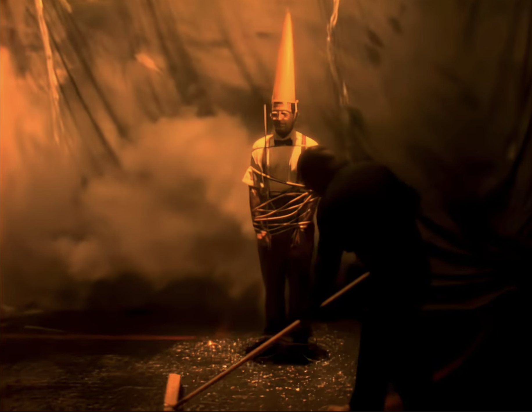 Authority figure tied up with a dunce hat on his head, while the janitor sweeps with a push broom.