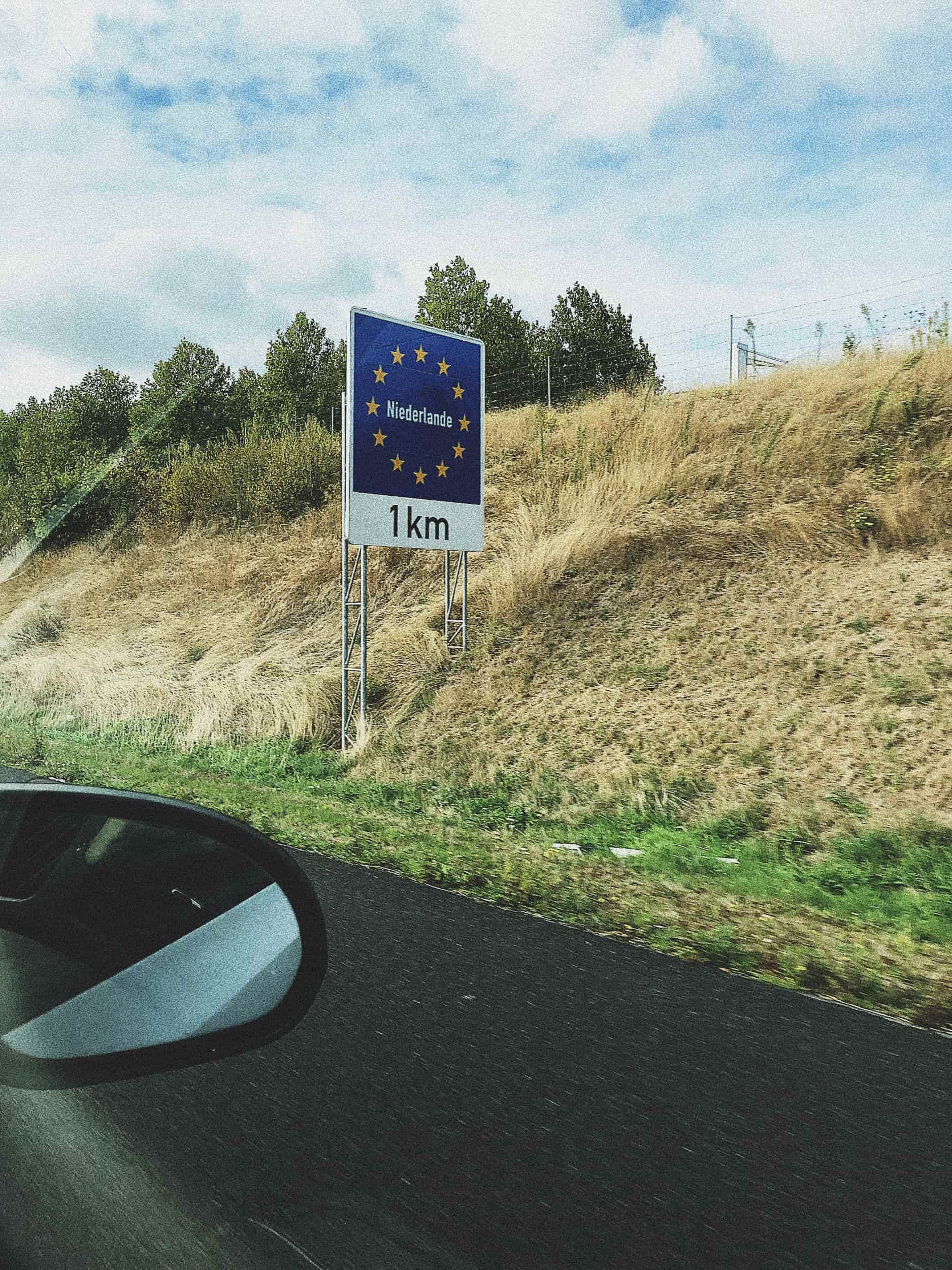 EU-KI-Souveraenitaet: Autobahnschild Niederlande 1km nahe ASML als Symbol europaeischer Technologie-Abhaengigkeit.