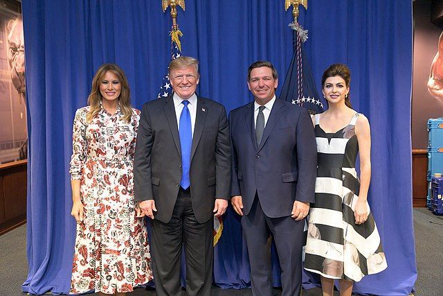 Ron & Casey DeSantis standing with Donald & Melania Trump, smiling at a camera in front of a blue background.
