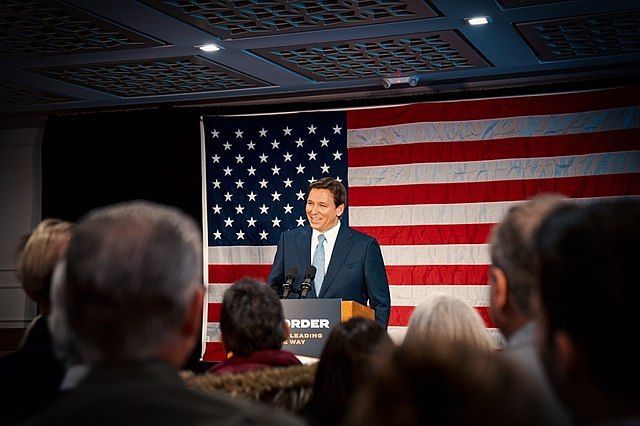 Ron DeSantis smiling and standing in behind a podium in front of an American crowd facing an audience. 