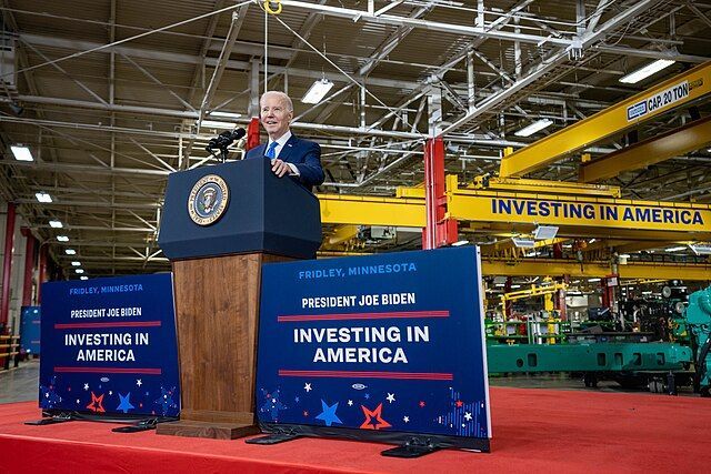 Joe Biden standing at a podium in a Minnesota factory with signs reading Investing in American on either side.