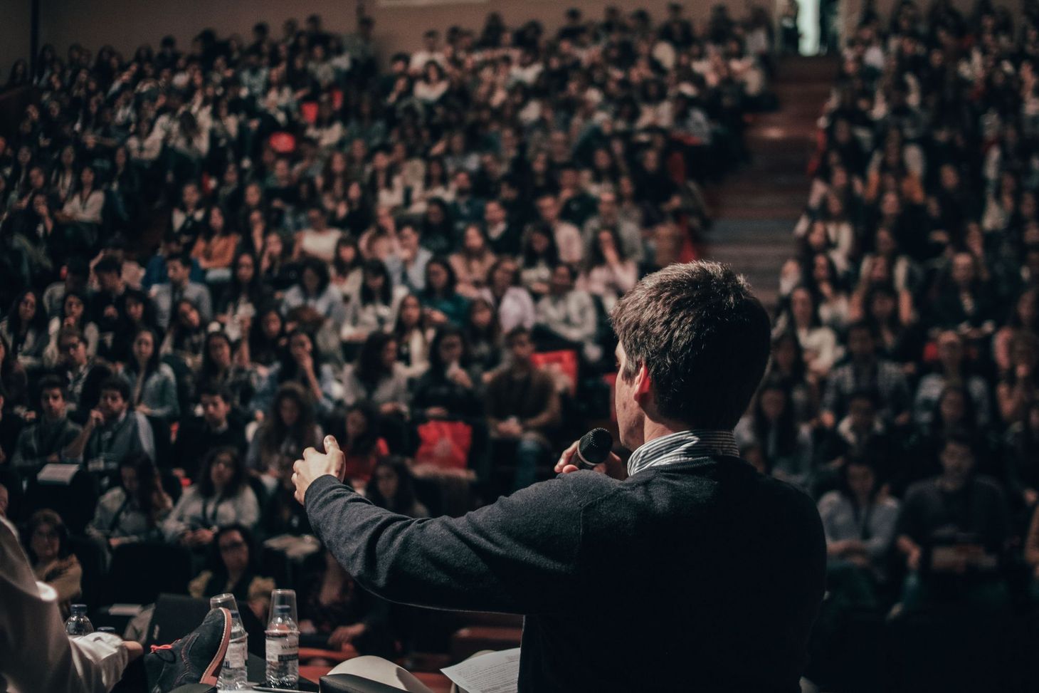 A man speaking before a crowd in a lecture hall, mic in hand, someone sitting next to him mostly out of frame. 