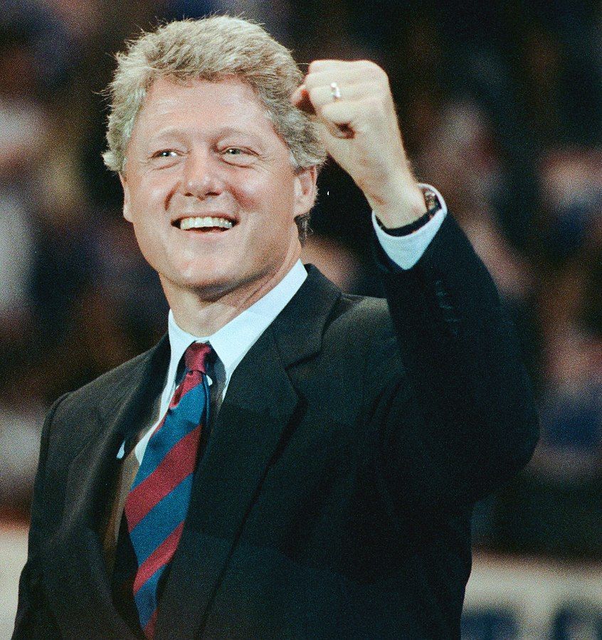 Bill Clinton smiling, hand raised in a cheering fist, at a 1992 campaign rally. 