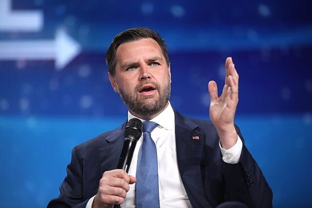 JD Vance seated and speaking, mic in hand, in front of a blue background. 