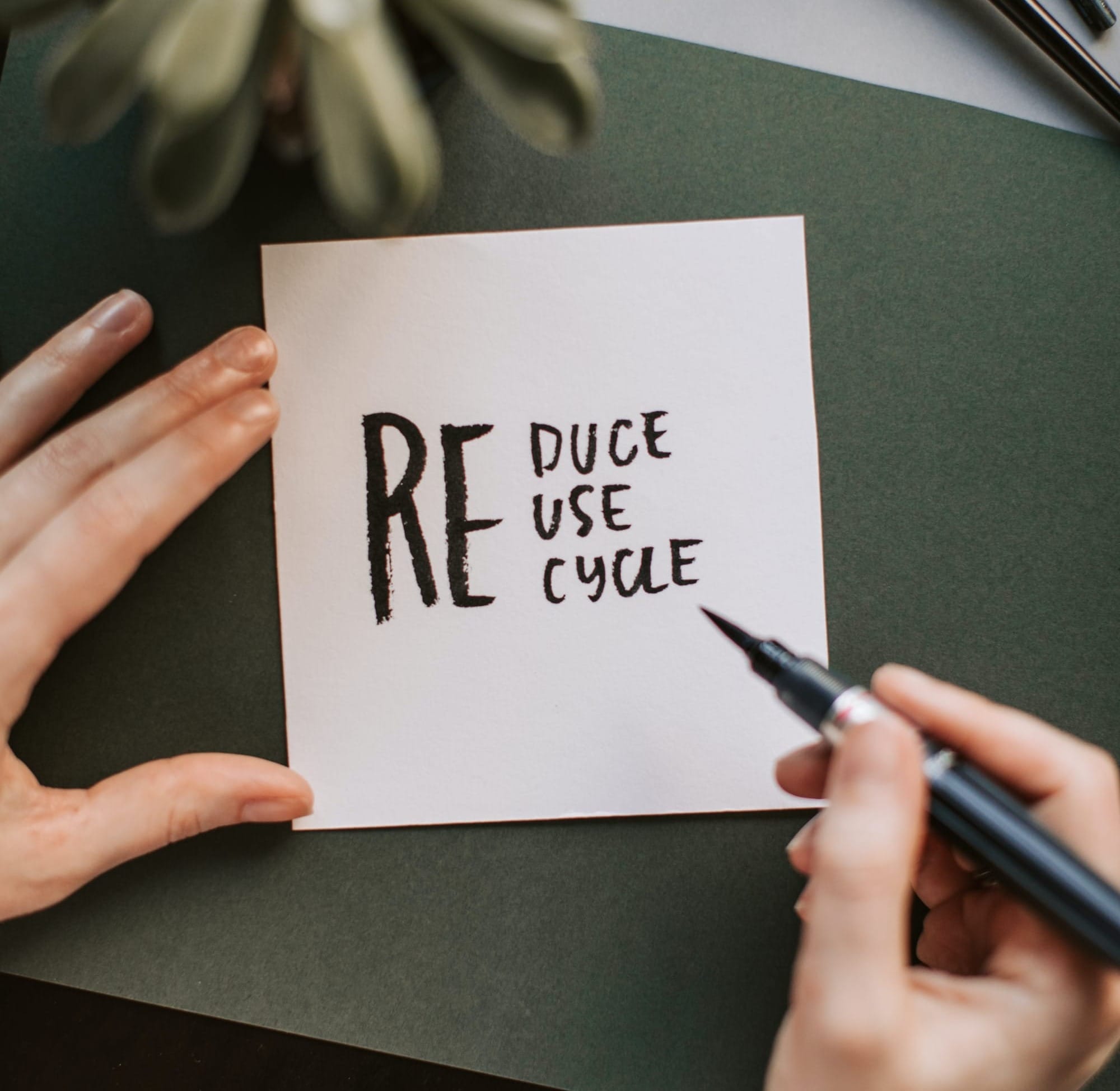 Hands writing, with black ink, on a paper with the words: reduce, reuse, recycle