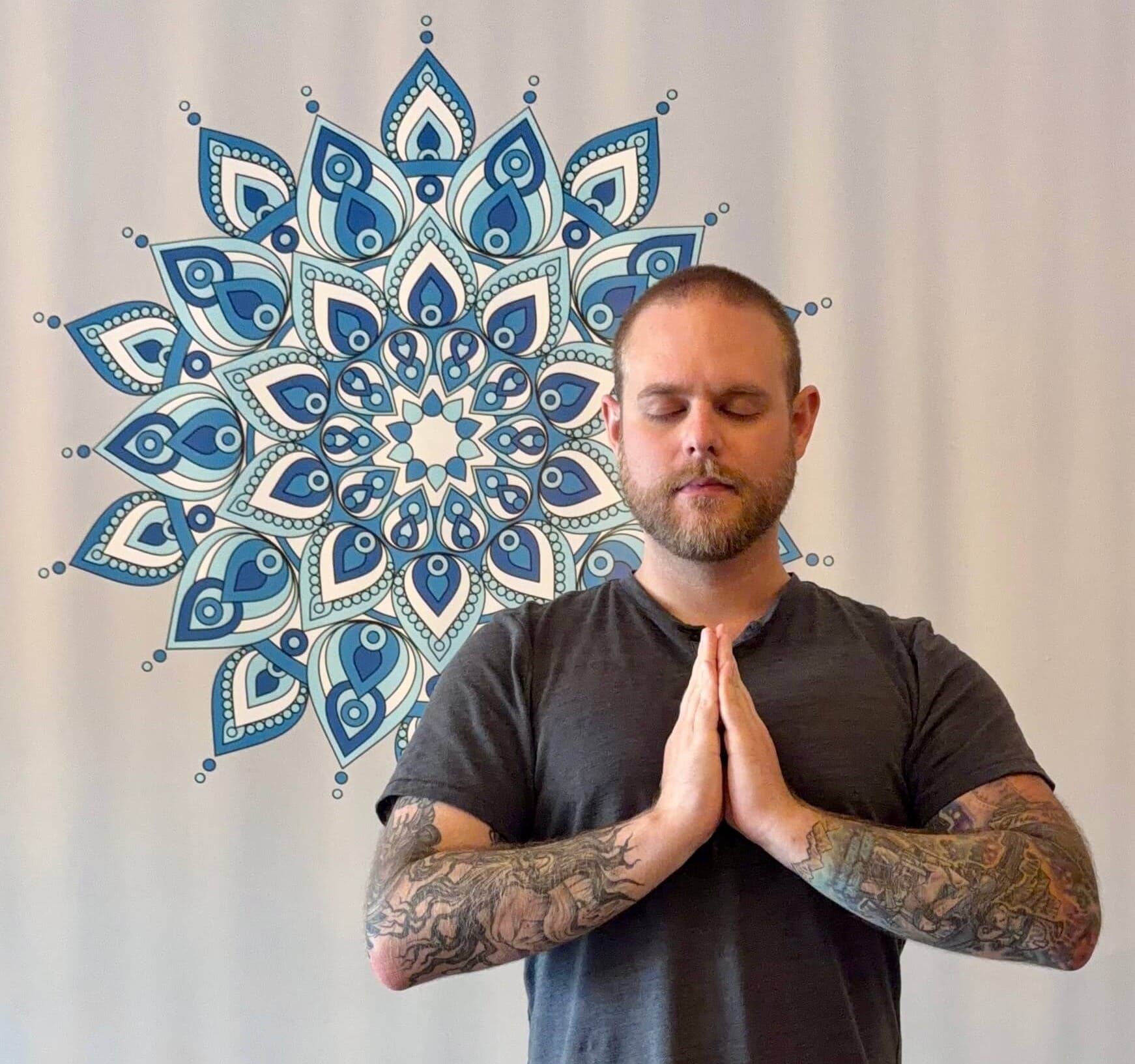 A man with tattooed arms stands with his palms together at his chest and eyes closed, in front of a blue mandala