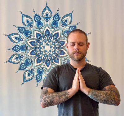 A man with tattooed arms stands with his palms together at his chest and eyes closed, in front of a blue mandala