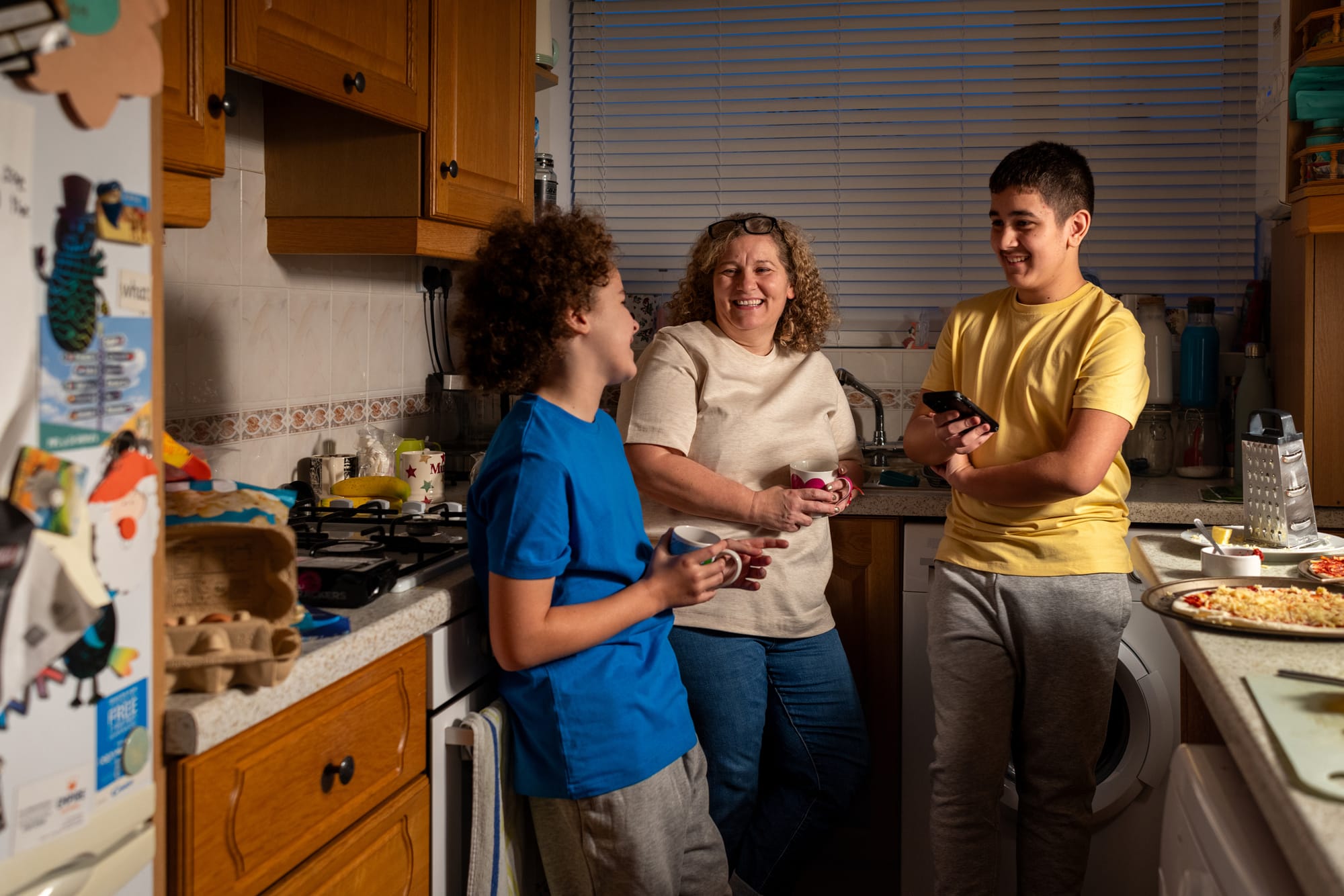 Two teenagers and a middle-aged adult stand in a kitchen, holding mugs and a mobile phone while talking. Wooden cupboards, kitchen appliances and a fridge covered with children's artwork can be seen around them.