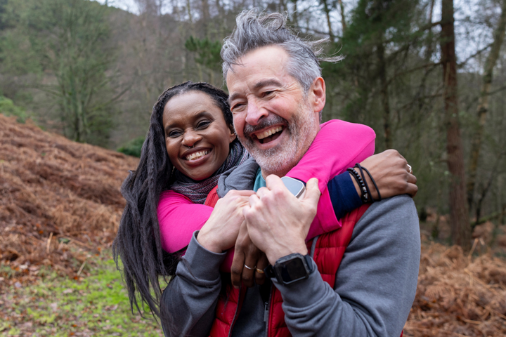 A man and woman hugging in a wooded clearing with smiles on their faces. 