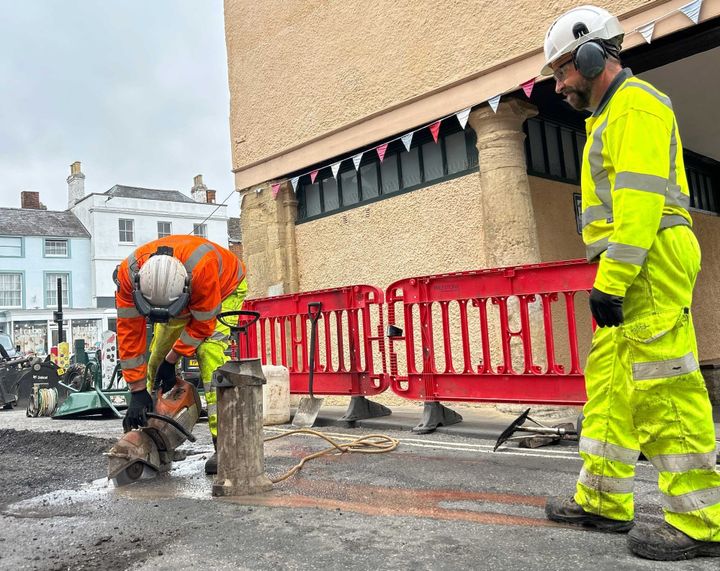 Two highways crew in protective gear fixing a damaged area of road - one male is bent over using a circular saw to cut into the tarmac. The other male looks on.