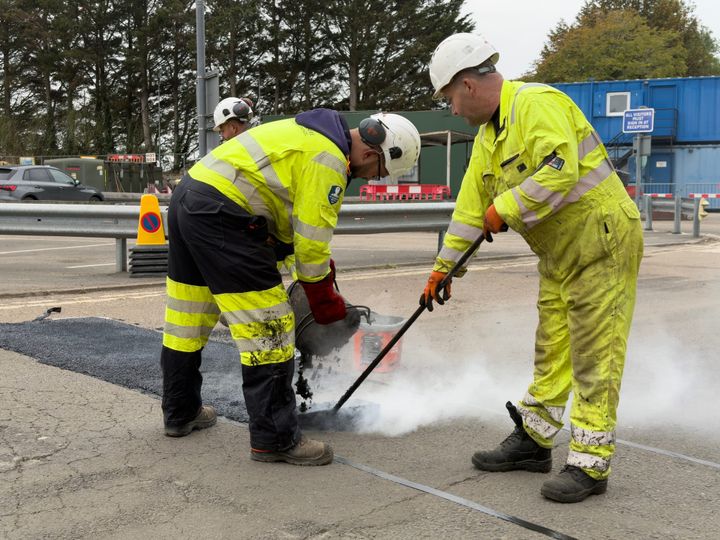 Two highways crew resurfacing an area of road. One male is bent over emptying tarmac while another make is spreading the tarmac.