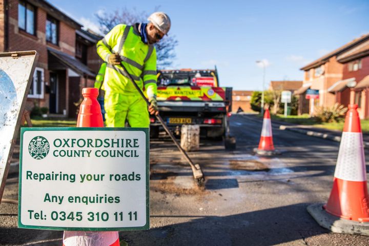 Highways worker in safety gear brushing over a repaired pothole in a residential street. He is surrounded by cones with a repairing your roads sign in the foreground with the enquiries number