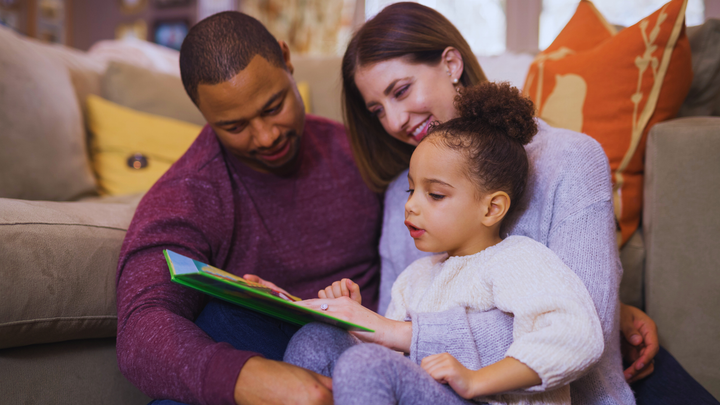 Young couple sat on the floor in their living room, a young girl is sat with them and they are all reading the same children' book