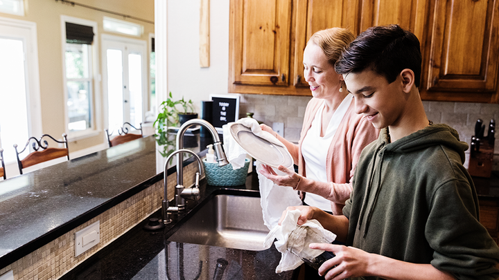 An adult and a teenager stand at a kitchen sink drying dishes together. The kitchen has wooden cupboards, a dark worktop and large windows letting in natural light. Plates and tea towels are 