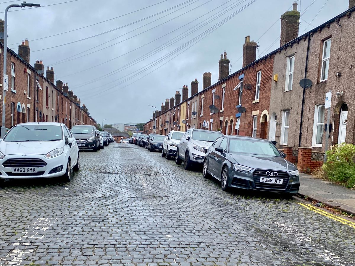 A cobbled road flanked with parked cars and terraced houses.