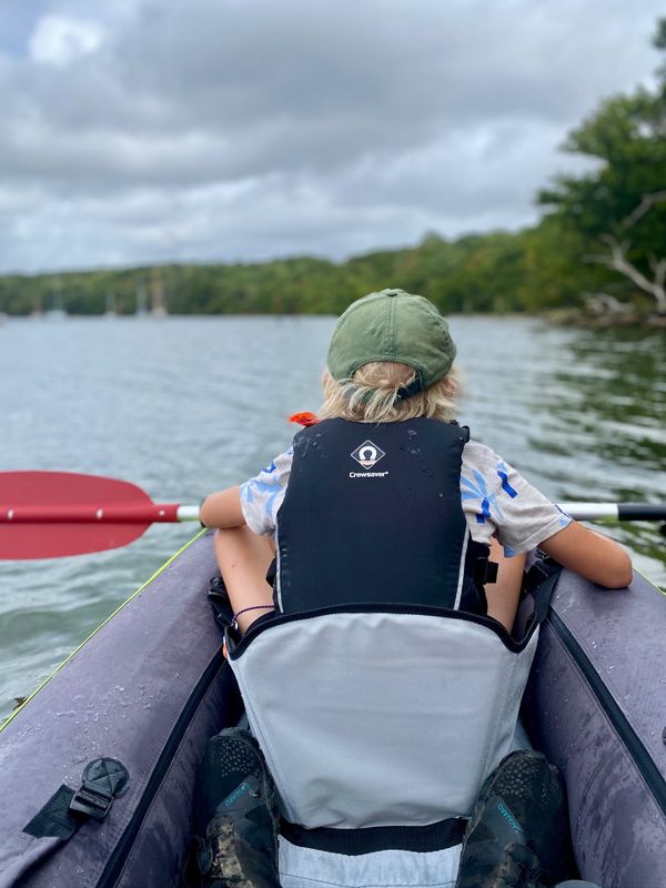 a boy at the front of an inflated kayak, photograph taken from the rear seat. River and trees beyond the kayak.