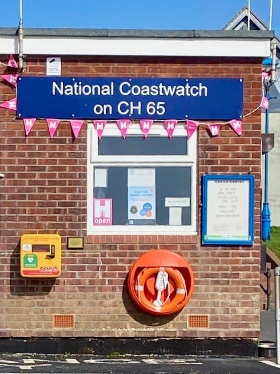 A brick building with mounted Defibrillator, Life ring and information board, hanging bunting and a sign reading ‘National Coastwatch on CH65’