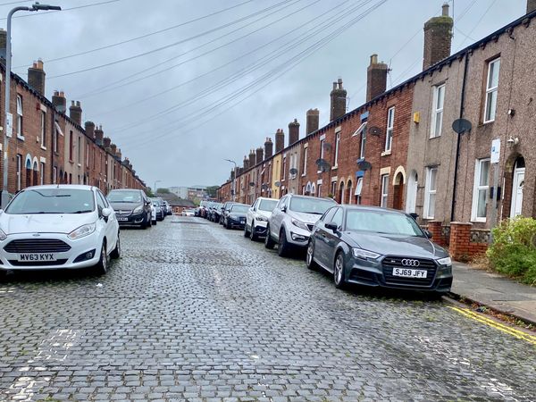 A cobbled road flanked with parked cars and terraced houses.