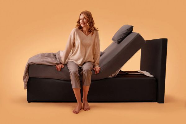 Frau sitzt auf einem Bett mit dem CosyLift, der die Matratze elektrisch in eine aufrechte Position anhebt